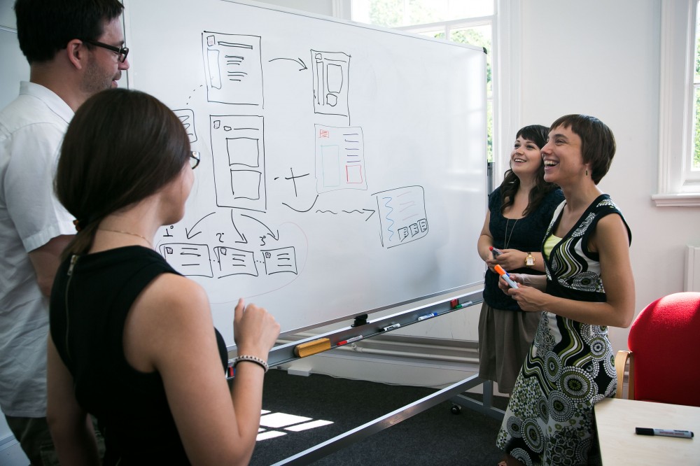 Designers standing around a whiteboard discussion some interfaces designs drawn on it.