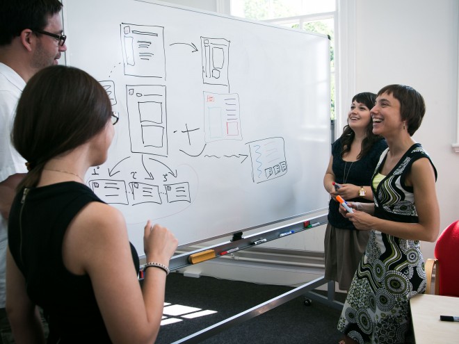 Designers standing around a whiteboard discussion some interfaces designs drawn on it.